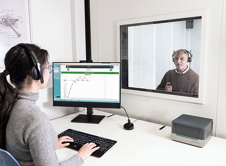 Clinician operating PC keyboard and ACT test screen in the Affinity Suite. The clinician has an Affinity Compact on their desk, and the clinician is looking through a glass window to a client sat on the other side in a booth. The client is wearing headphones and pushing a patient response button.