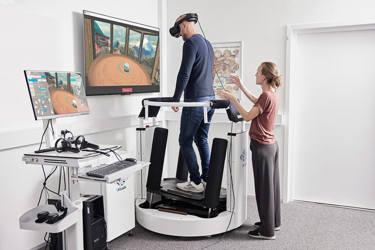 A patient is standing on the enclosed dynamic force plate, which is tilting backward to the right. A clinician is standing behind the patient on the floor at a short distance, with their right hand held up to be ready to support the patient should they fall.