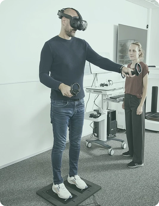 Patient wearing Vive Focus Vison VR headset performing a training module with controllers while stood on the Virtualis StaticVR force plates with a clinician observing