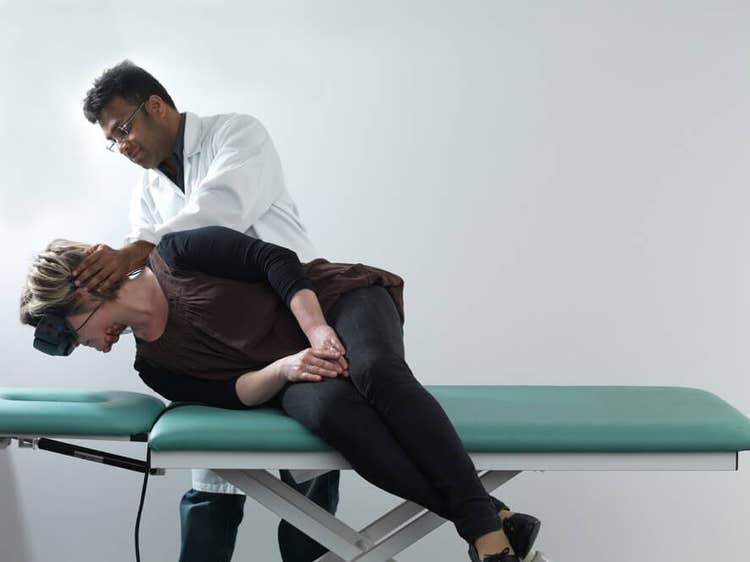 Female patient in black clothes lying on her right on examination table with her legs dangling off the edge of the table. Male clinician stood behind is turning her head 45 degrees toward the right.