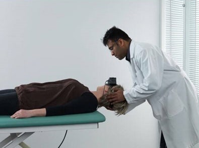 Clinician stood behind patient lying on their back on a green examination bed. The clinician is turning the patient’s head toward the right at a 45-degree angle.
