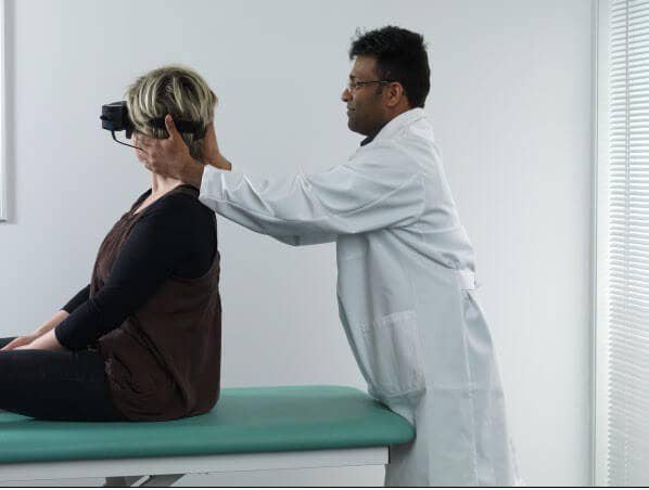 Female patient sitting on examination table with back toward a male clinician. The clinician is turning the patient's head 45 degrees toward the right.