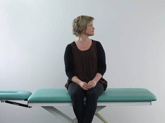 Woman in black clothes sitting on examination table with legs dangling off the edge of the table. She has her head turned about 90 degrees toward the left.