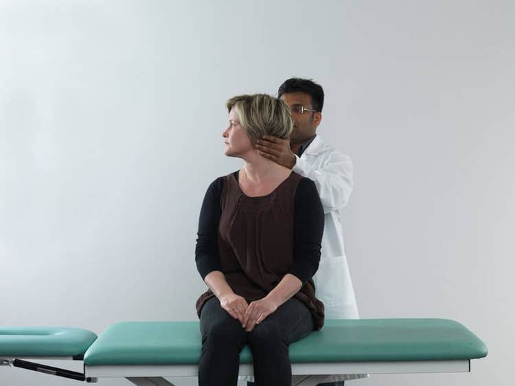 Female patient in black clothes sitting on examination table with legs dangling off the edge of the table. Male clinician stood behind the patient, and is turning her head about 90 degrees toward the right.