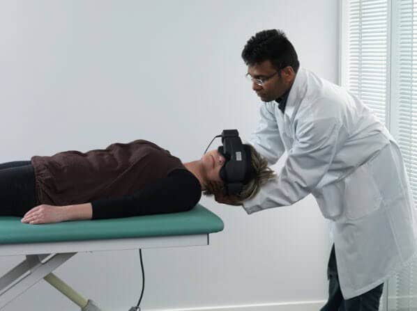 Female patient lying on her back on examination table, with head hanging over the edge of the table. Male clinician is turning the patient's head toward the left.