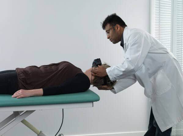 Female patient lying on her back on examination table, with head hanging over the edge of the table. Male clinician is turning the patient's head toward the right.