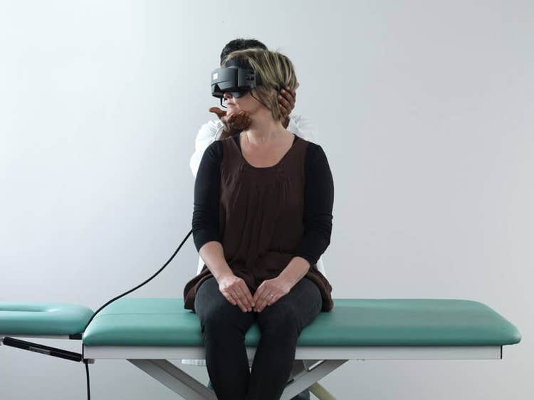 Female patient in black clothes sat on examination table with her legs dangling off the edge of the table. Male clinician stood behind is turning her head 45 degrees toward the right.