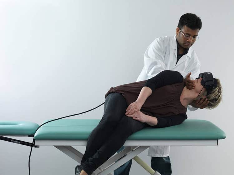 Female patient in black clothes lying on her left on examination table with her legs dangling off the edge of the table. Male clinician stood behind is turning her head 45 degrees toward the right.