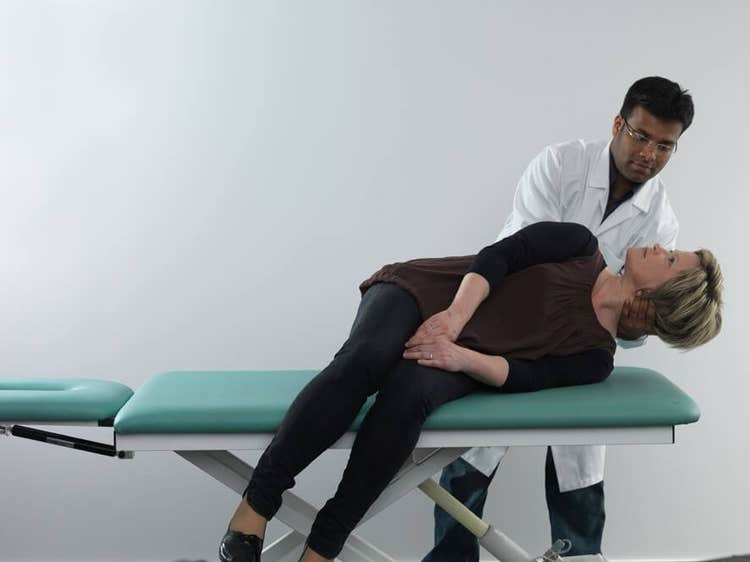 Female patient in black clothes lying on her left side on examination table with legs dangling off the edge of the table. Male clinician stood behind the patient, and is turning her head about 90 degrees toward the right.