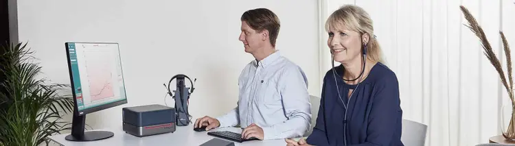 Clinician at a fitting screen performing real-ear measurement while the client sits beside the desk wearing probe tubes