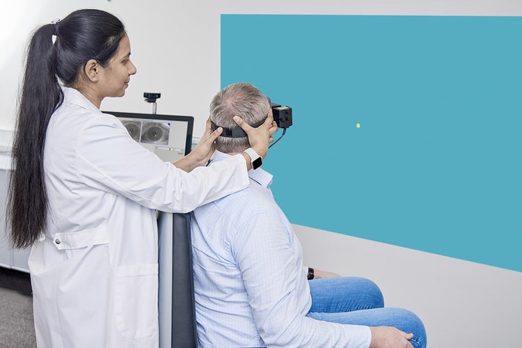 Clinician performing Gaze test in a patient. The patient is sat in the Orion Reclining rotary chair and fixated with VisualEyes VNG goggles, looking toward a dot projected onto a wall. Their body is facing 45 degrees to the right of the stimulus, and the clinician is turning the patients head 45 degrees to the left to look at the stimulus. The positioning is the same for the Smooth Pursuit Neck Torsion test.
