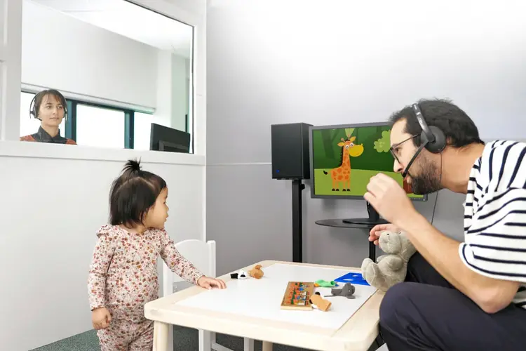 An audiologist is looking into a booth with a second audiologist who is doing VRA testing with a child