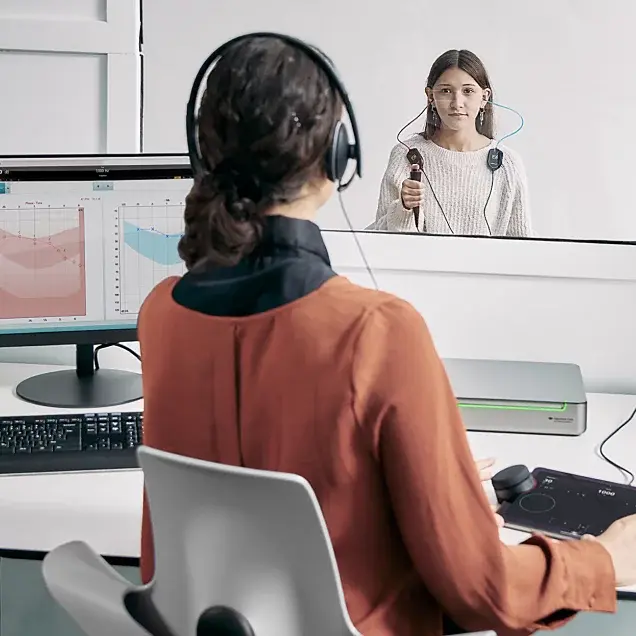 A clinician using the Equinox Evo and Touch Keyboard to perform audiometry in a teenager sitting in a booth