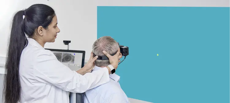 Rear view of patient wearing VNG goggles. A clinician stood behind the patient is turning the patient's head 45 degrees to the left, with the patient looking toward a dot stimulus projected on to the wall in front of them