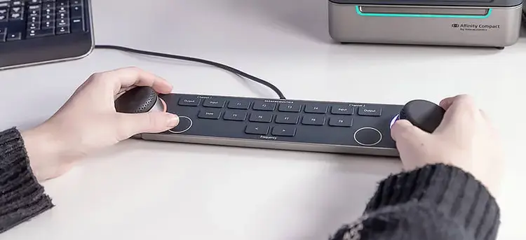 Audiometer Keyboard on a white desk in front of a PC keyboard and the Affinity Compact. A person is operating the keyboard, with their hands placed on the two dials located on the left and right sides of the keyboard. There are several keys between the multi-functional dials, which include and are not limited to input and output for channels one and two, store, two keys to decrease or increase the intensity, and nine F-keys labeled F1 to F9.